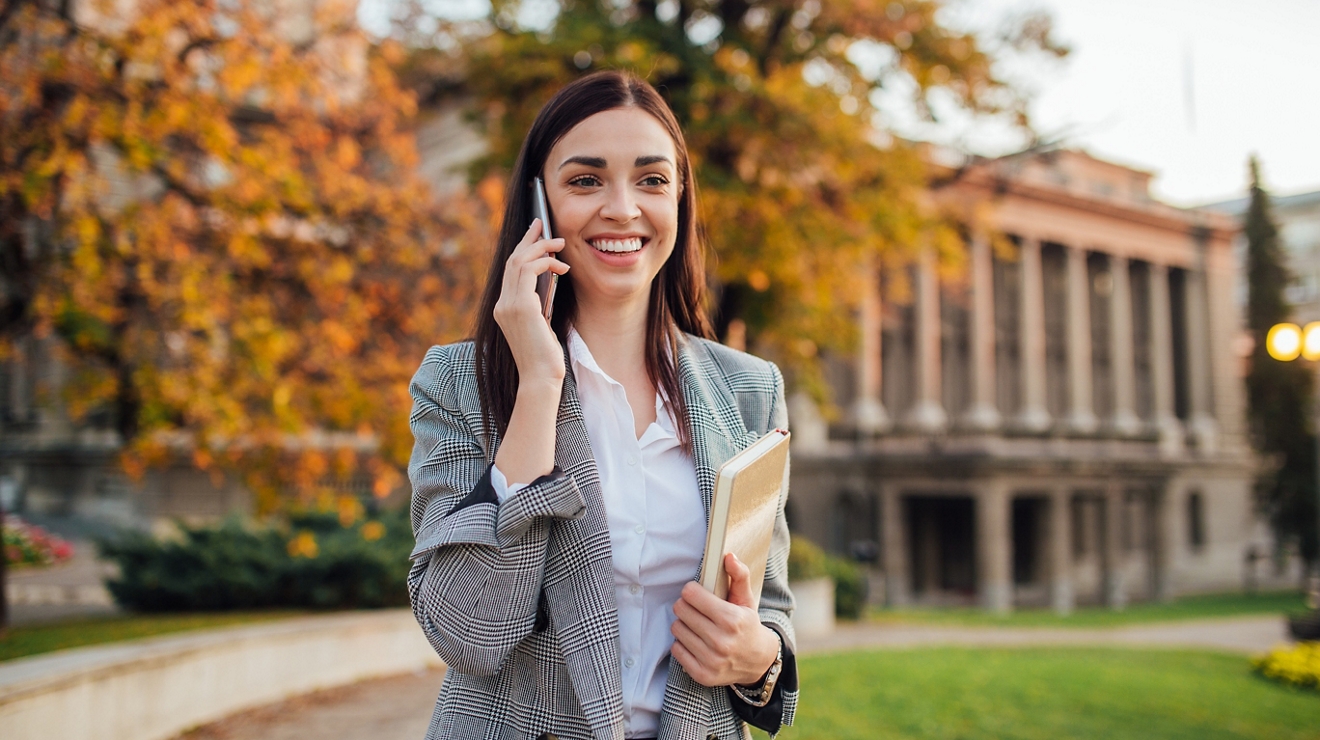 Joven mujer luce feliz mientras habla por teléfono junto al edificio de la universidad.