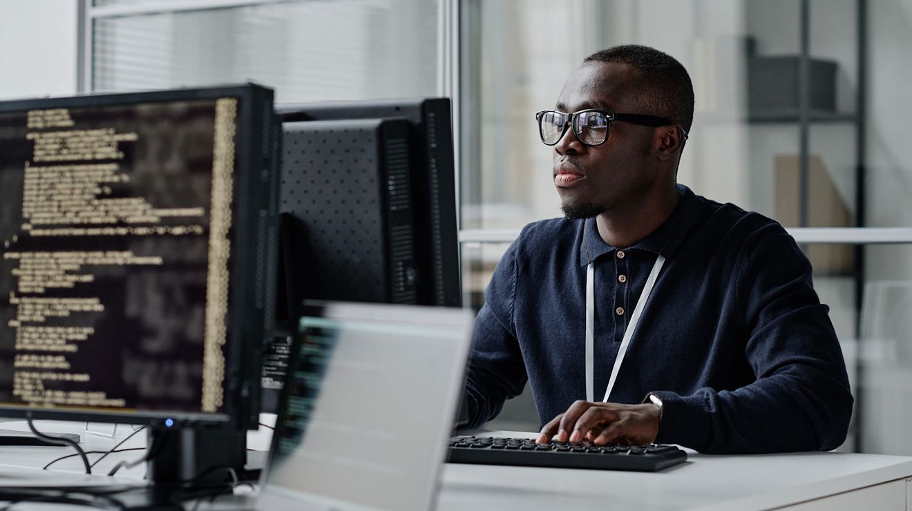 Joven desarrollador afroamericano con anteojos concentrado en su trabajo por Internet en la computadora, sentado en su lugar de trabajo.