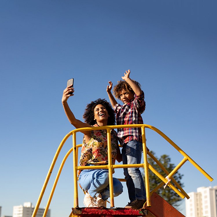 Mujer tomándose una selfie con un niño con los brazos levantados sobre un juego de parque infantil.