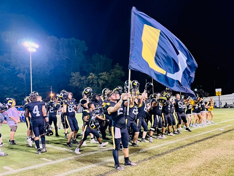 Jugadores de fútbol americano de Union High School, vestidos con uniformes azules y amarillos, se reúnen en el campo y ondean una bandera de Union High School.
