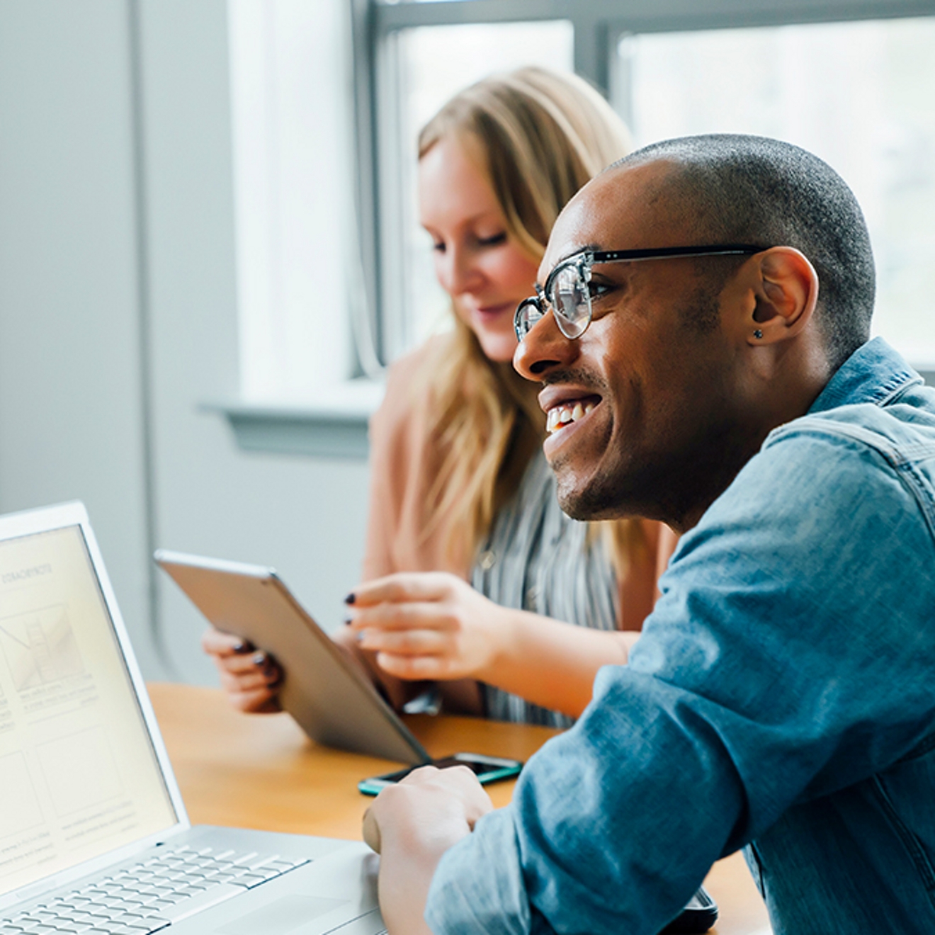 Dos compañeros de trabajo interactuando con una laptop, una tablet y un teléfono.